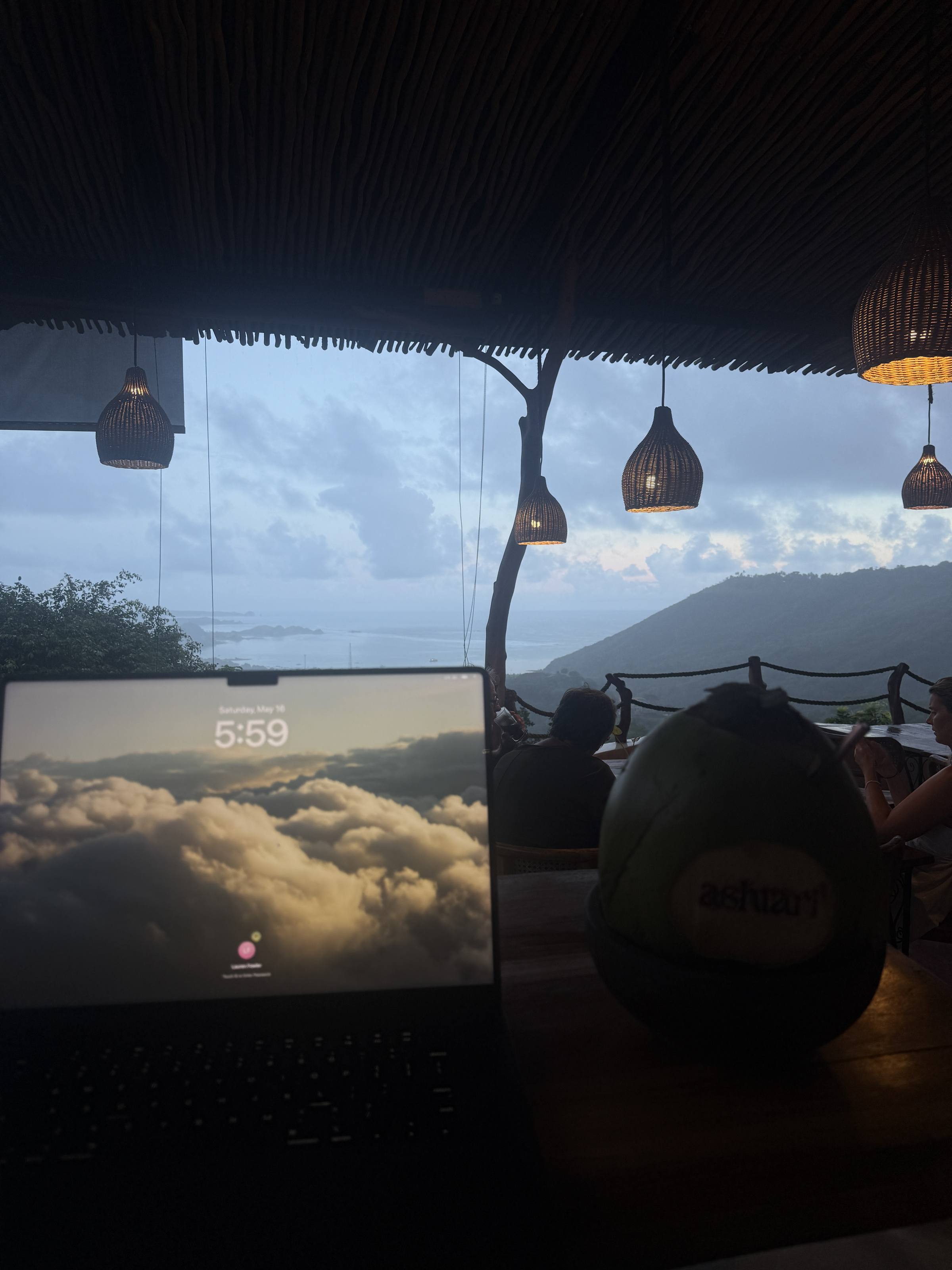A laptop on a cliff-edge café table at dusk, lanterns above and cloud below