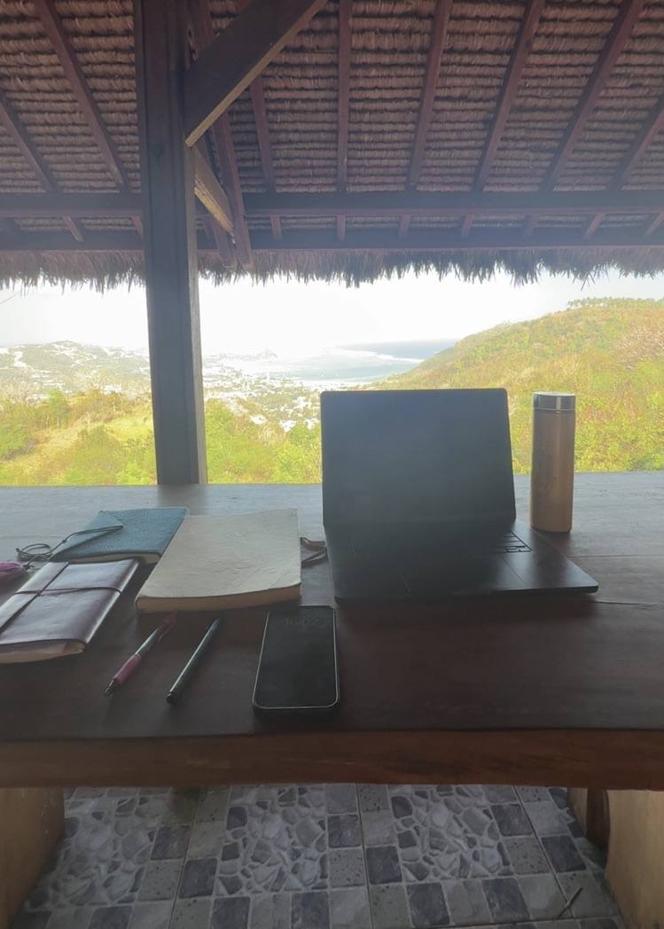 A laptop and papers on a desk under a thatched roof facing the hills