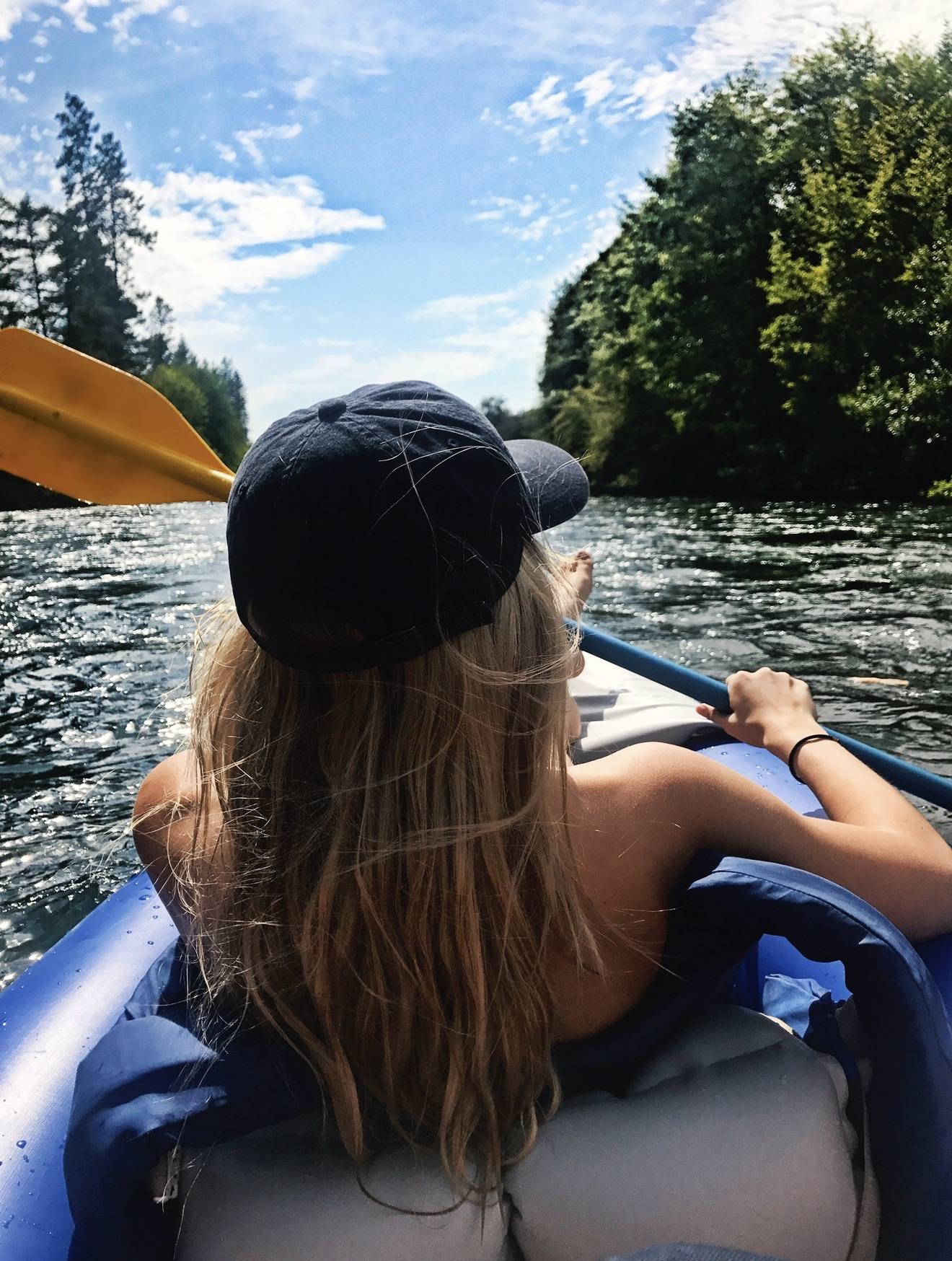 Paddling an inflatable kayak down the Rogue River in a black cap, firs on both banks